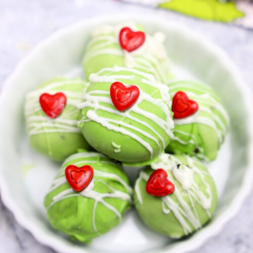 A delightful plate of green truffles, reminiscent of festive Grinch Oreo Balls, is topped with a white drizzle and charming red heart decorations. Perfect for Christmas recipes that add a touch of whimsy to your holiday celebrations.