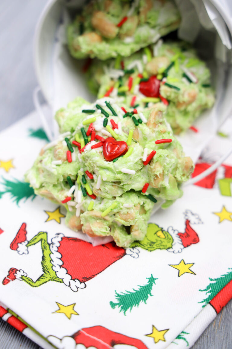 Green candy clusters with colorful sprinkles and heart decorations, all set on a festive napkin featuring a cartoon character in a Santa suit. These whimsical treats, dubbed Grinch Poop Peanut Clusters, add a fun and playful touch to your holiday celebrations.