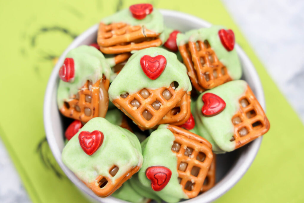 Indulge in these Grinch Heart Pretzel Bites—a bowl of pretzels partially dipped in green icing and adorned with playful red heart-shaped candies.