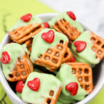Bowl of Grinch Pretzel Bites, each square pretzel partially coated in green icing and topped with a small red heart-shaped candy.