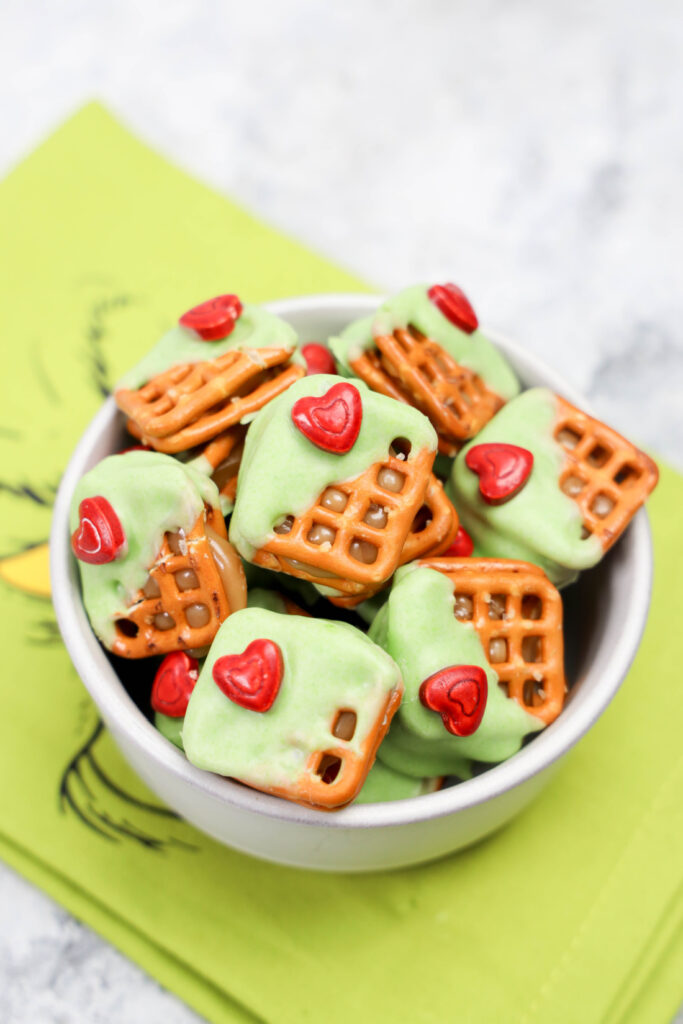 A bowl of Grinch Heart Pretzel Bites, where pretzel squares are partially dipped in green icing and adorned with a small red heart, all presented on a festive green napkin.