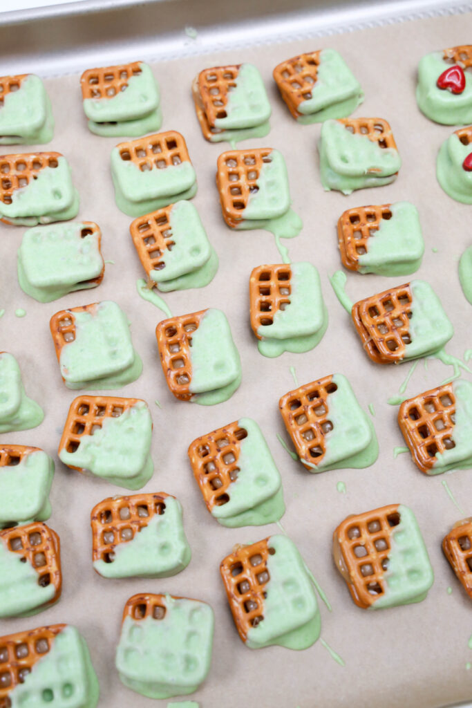 Grinch Heart Pretzel Bites, partially dipped in light green coating, are elegantly arranged on parchment paper. Some showcase small red heart sprinkles adding a charming touch.