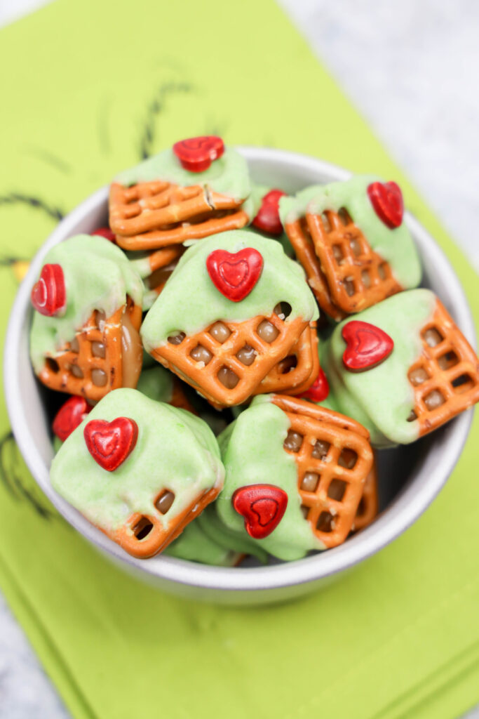 A bowl of Grinch Heart Pretzel Bites partially dipped in green frosting, each topped with a small red heart candy, sits on a festive green napkin.
