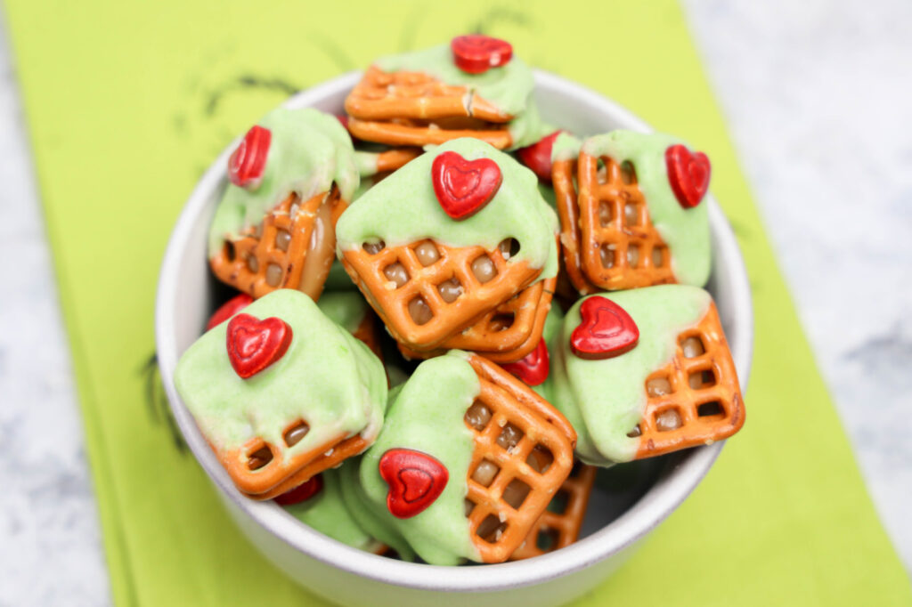 A bowl of Grinch Heart Pretzel Bites features square pretzels partially coated in green icing, each topped with a red heart decoration.