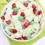 A bowl of festive Grinch Puppy Chow snack mix, featuring green and white hues combined with playful red heart-shaped candies, rests on a green cloth adorned with whimsical cartoon eyes.