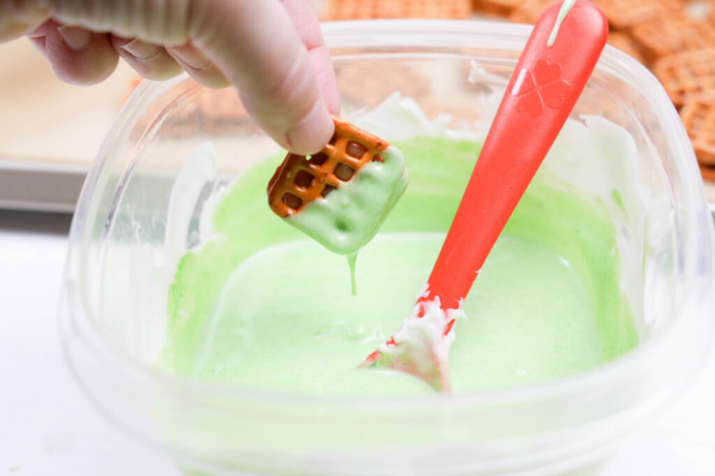 A hand dips a pretzel square into green frosting in a plastic container, using a red spatula, creating delightful Grinch Heart Pretzel Bites.