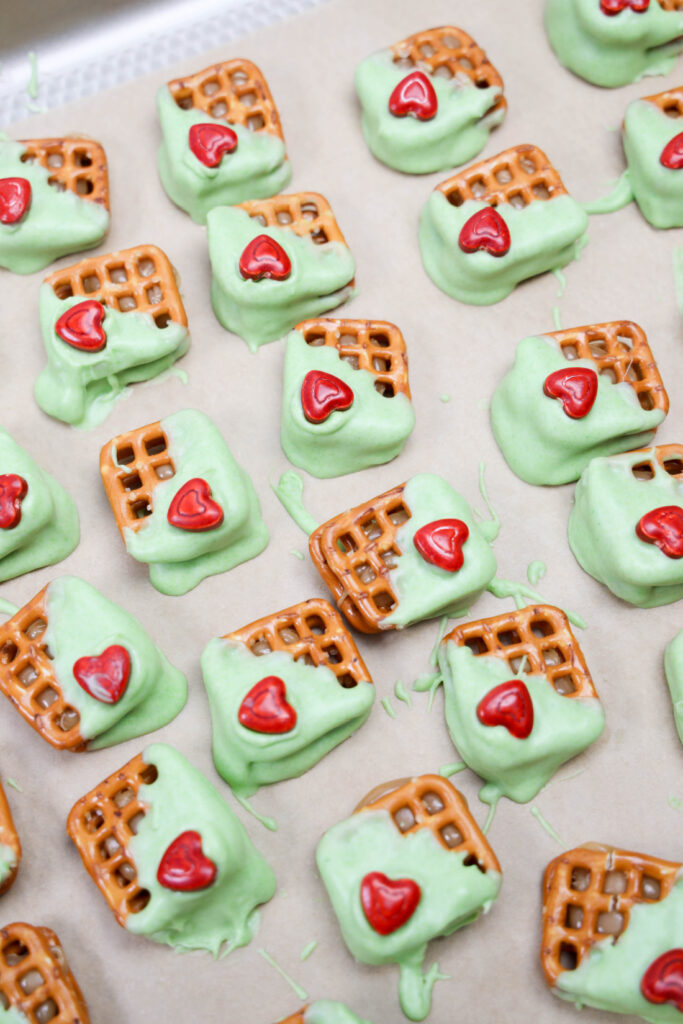 Grinch Heart Pretzel Bites: Pretzels topped with light green frosting and red heart-shaped candies are carefully arranged on parchment paper.