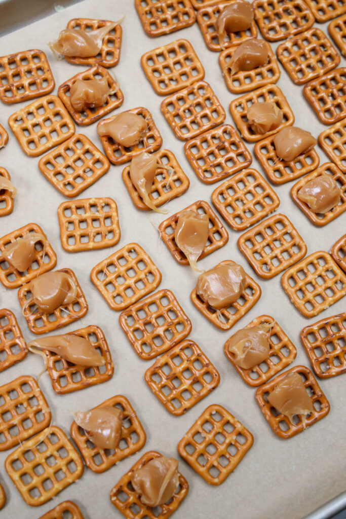 Square pretzels topped with dollops of caramel placed on a parchment-lined baking sheet, these Grinch Pretzel Bites are the perfect festive treat.