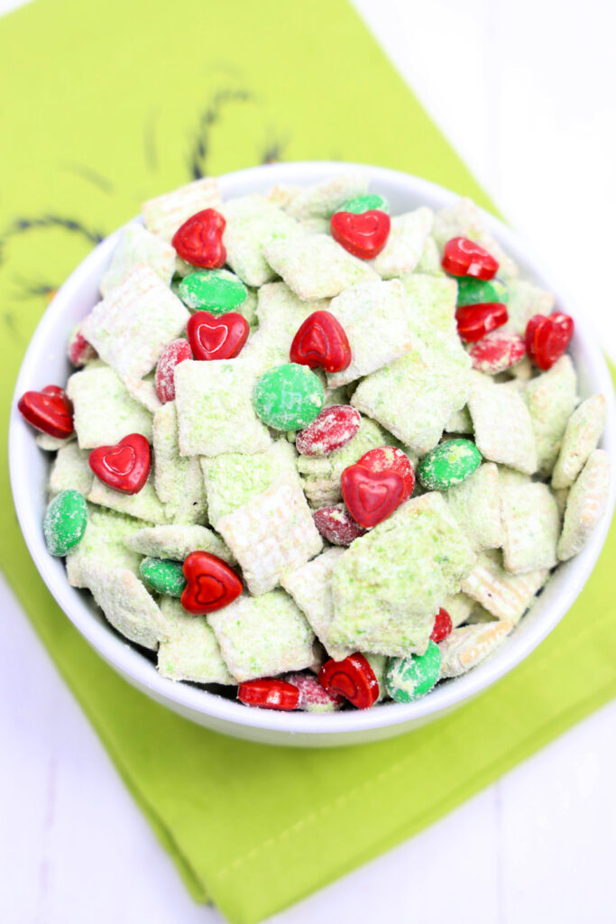 A bowl of Grinch Puppy Chow, featuring white chocolate-covered cereal mix, sprinkled with small red heart candies and vibrant green circular candies, all elegantly placed on a green napkin.