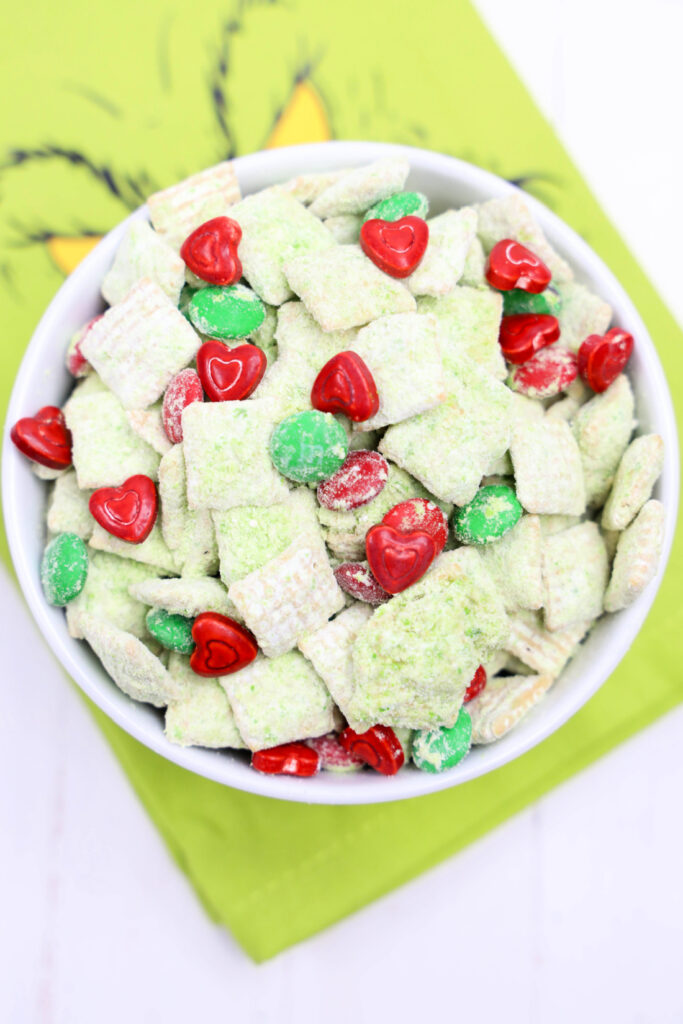 A bowl of Grinch Puppy Chow with green and white snack mix adorned with red heart-shaped candies sits on a green cloth.