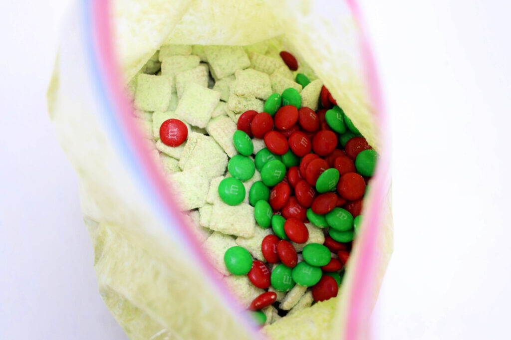 Open a bag of Grinch Puppy Chow, featuring cereal squares covered in powdered sugar and dotted with red and green chocolate candies.