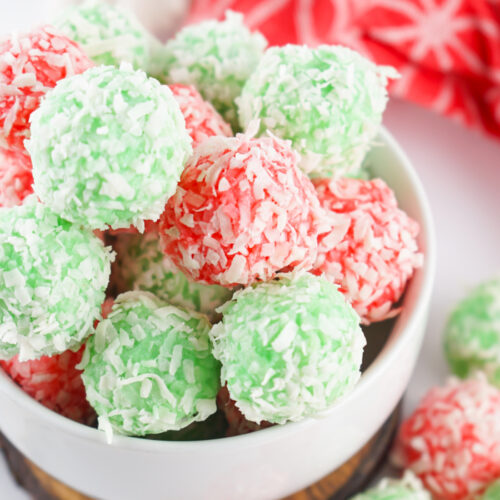 A bowl of red and green jello balls, coconut-covered and perfect as easy holiday treats, sits against a red patterned cloth in the background.