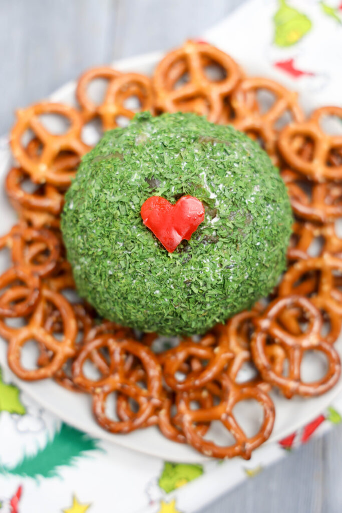 A Grinch cheese ball, tinted green and topped with a small red heart, sits charmingly amidst a ring of crunchy pretzels on a plate.