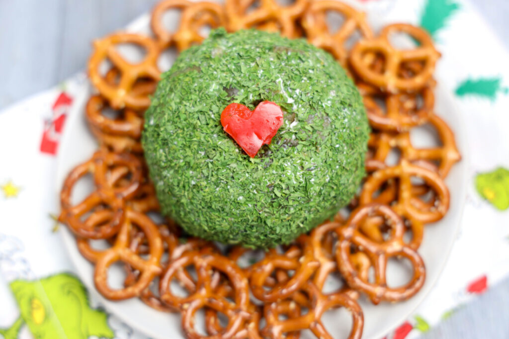A Grinch Cheese Ball, adorned with a red heart, sits on a white plate surrounded by pretzels.