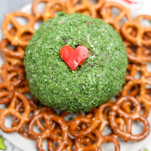 A Grinch Cheese Ball, adorned with a small red heart on top, sits at the center of a white plate, surrounded by crunchy pretzels.