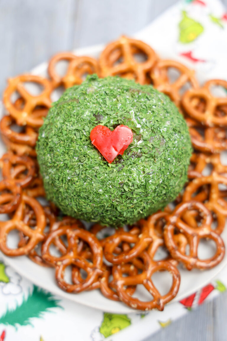 A Grinch Cheese Ball, vibrant green and adorned with a red heart-shaped decoration, sits elegantly on a white plate, surrounded by crunchy pretzels.