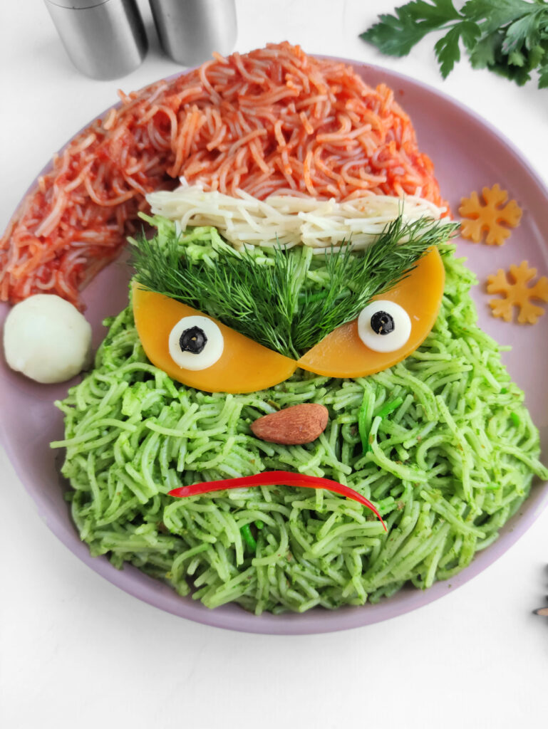 A plate of Grinch vermicelli pasta is artistically arranged to resemble the character, featuring green pasta for the face, a red hat, and garnishes forming expressive eyes and a mischievous mouth.