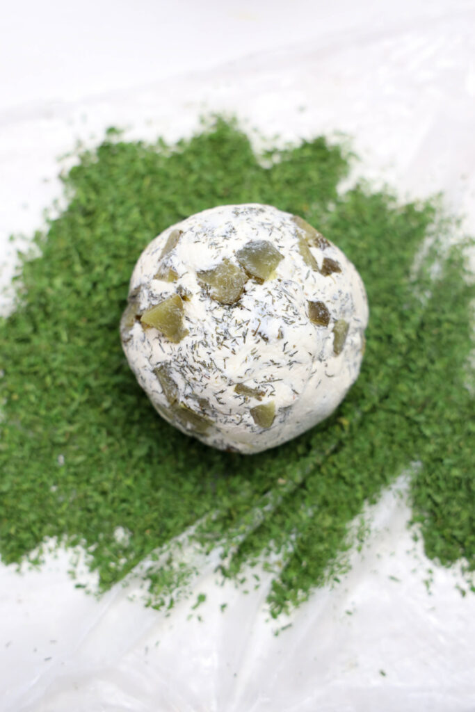 A Grinch Cheese Ball, adorned with herbs and what appear to be diced pieces of pickles, is nestled on a lush bed of chopped parsley.