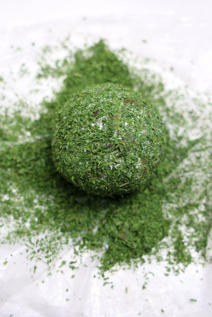 A Grinch Cheese Ball sits as a round object encrusted with green herbs, resting on a white surface scattered with even more herbs.
