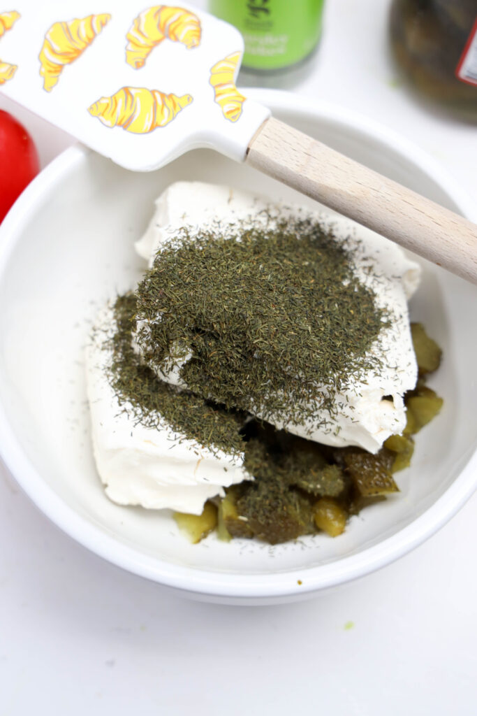 A bowl brimming with Grinch Cheese Ball goodness: cream cheese, chopped pickles, and a heap of dried dill. A spatula adorned with a whimsical croissant design rests on the edge.