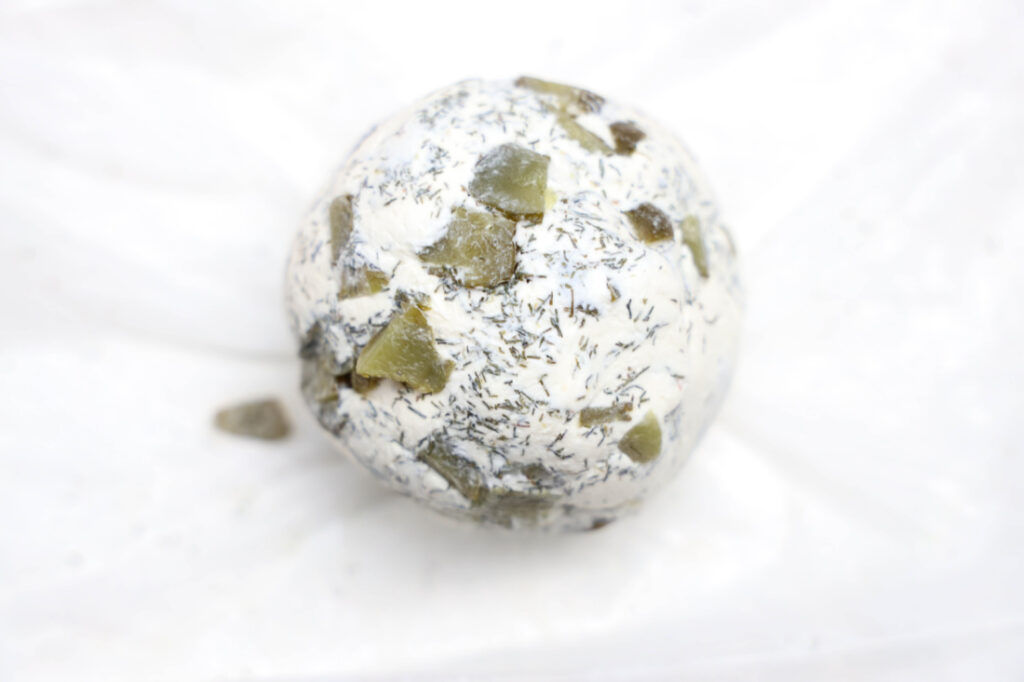 A Grinch Cheese Ball sits on a white background, adorned with herbs and small chopped pickles, offering a festive twist to the classic cheesy delight.
