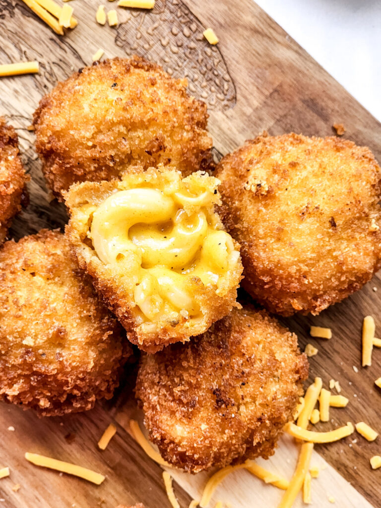 Close-up of crispy fried mac and cheese bites on a wooden surface, with one split open to reveal the gooey pasta inside, surrounded by scattered shredded cheese.