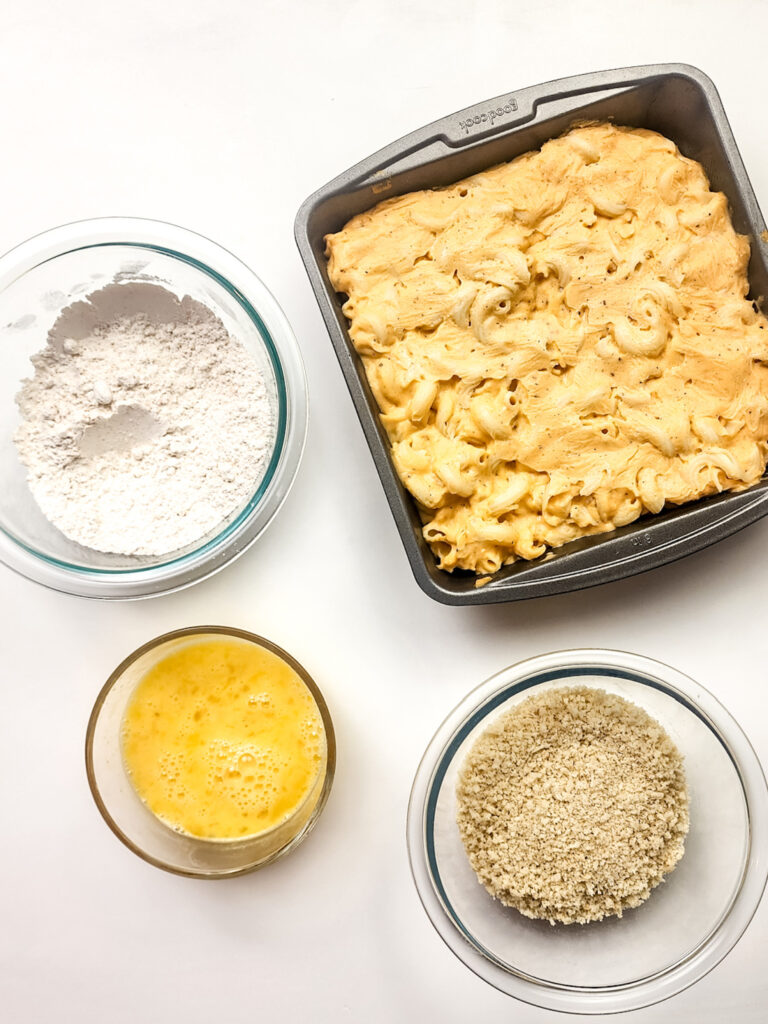 A square pan of baked macaroni and cheese rests beside bowls of flour, beaten eggs, and breadcrumbs on a white surface, ready to transform into crispy fried mac and cheese bites.