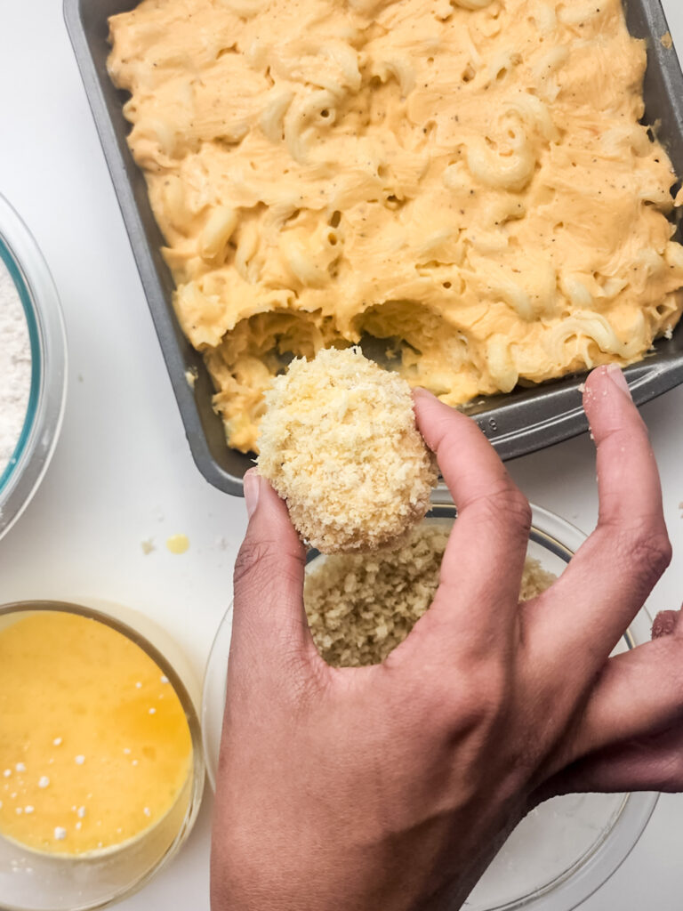 A person coats a ball of mac and cheese in breadcrumbs over a bowl, crafting deep-fried mac and cheese bites. Nearby are a baking dish of mac and cheese, a bowl of flour, and a bowl of beaten eggs.