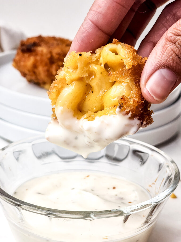 A hand holding a crispy fried mac and cheese bite dipped in creamy sauce, with a plate of deep-fried mac and cheese bites in the background.