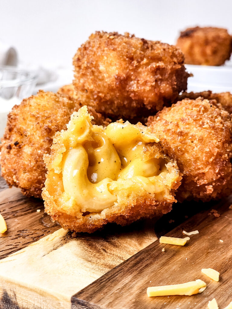 Close-up of crispy Fried Mac and Cheese Bites on a wooden surface, one cut open revealing creamy macaroni inside. Shredded cheese is scattered nearby.