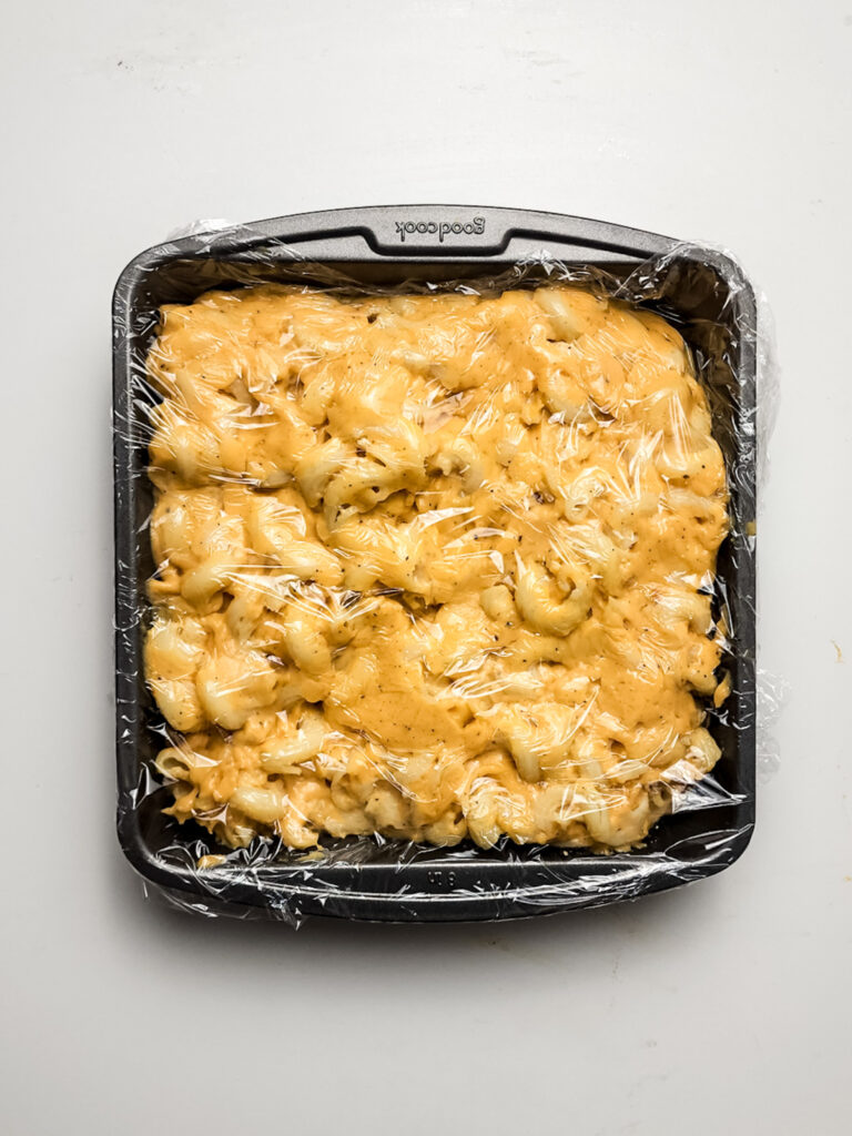 A black baking dish sits on a white background, holding macaroni and cheese covered with plastic wrap, reminiscent of homemade Mac and Cheese Bites in their pre-fried glory.