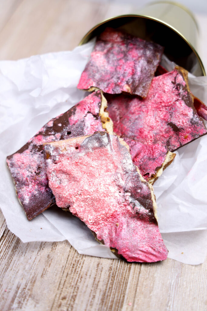 Glitter Valentine's Cracker Candy: A Sweet & Sparkly Treat 11 Pieces of pink and brown chocolate bark, reminiscent of Valentine's Cracker Candy, spill from a metal container onto parchment paper on a wooden surface.