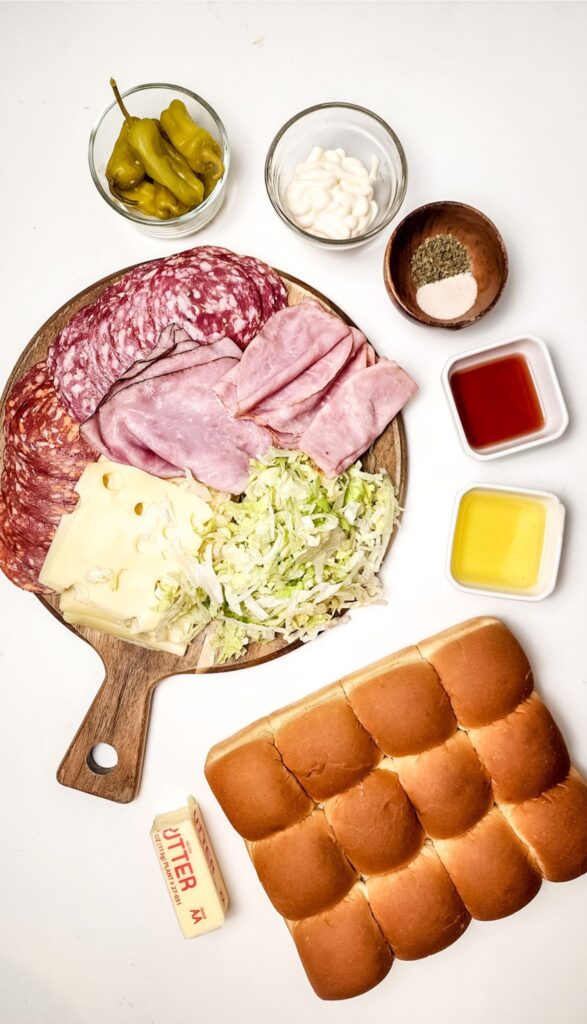 Easy Italian Sliders Recipe 2 Ingredients for Italian sliders on a board: sliced meats, cheese, shredded lettuce, and condiments. Bread rolls, butter, and small bowls of seasonings and oils are arranged around. Perfect for crafting an easy Italian sliders recipe that's both delicious and simple to prepare.