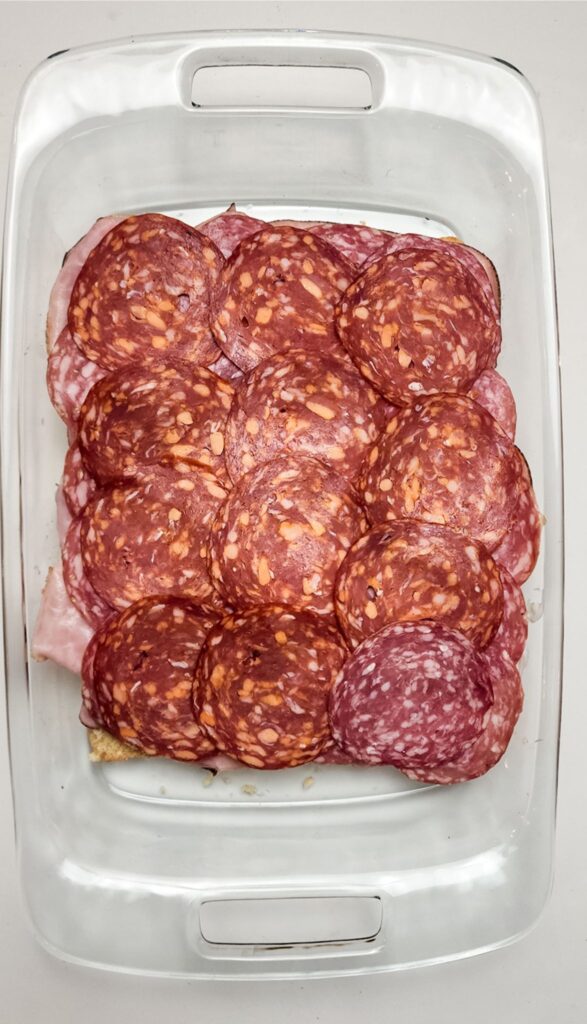 Easy Italian Sliders Recipe 4 Rectangle glass dish with overlapping slices of salami and ham covering layered food beneath, creating a tempting base for these easy Italian sliders.