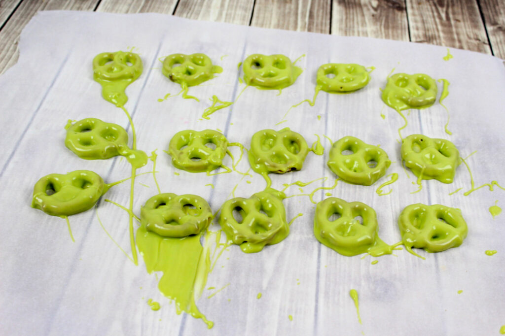 Pretzels coated in green chocolate are laid out on a parchment-lined surface, with some melted chocolate drizzled around.