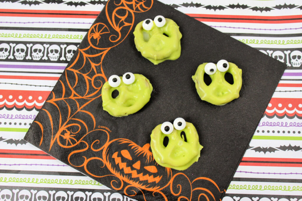 Four green monster-faced cookies with googly eyes on a Halloween-themed napkin.