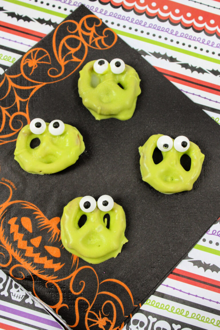 Four green monster face pretzels with candy eyes on a black Halloween-themed napkin.
