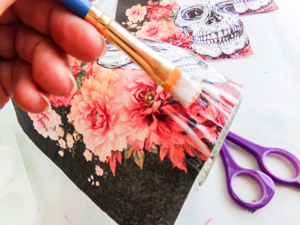 A hand applies glue with a brush to paper featuring skull and flower designs. Scissors are visible in the background.