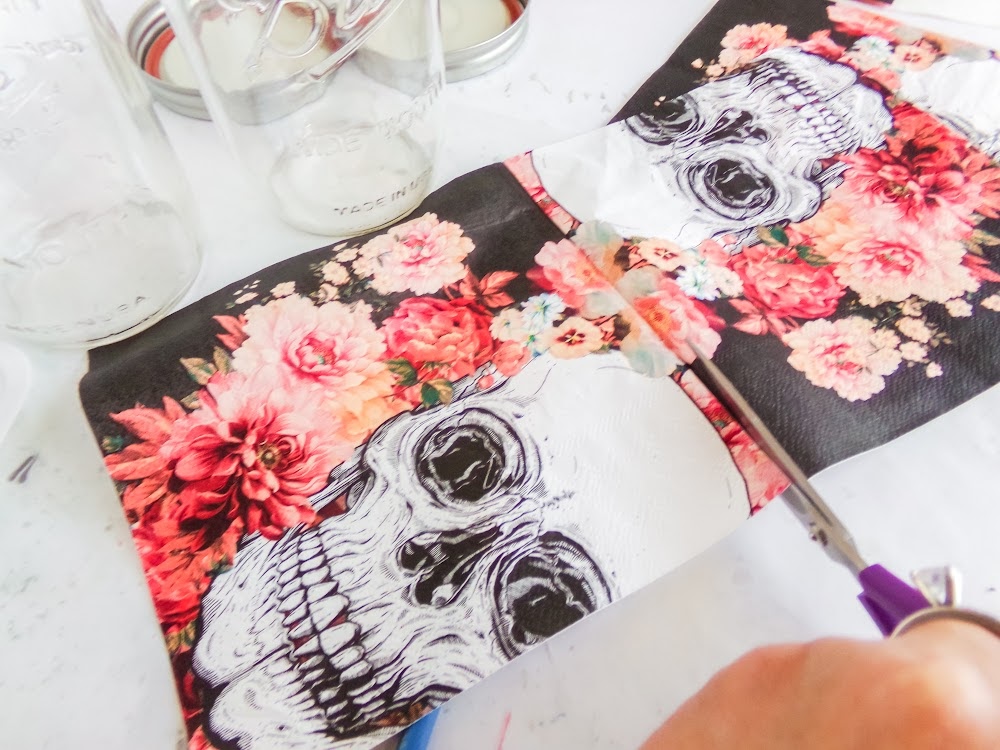 A person cuts floral skull-patterned paper next to glass jars on a white surface.