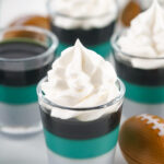 Layered dessert shooters topped with whipped cream, featuring teal and black jelly layers in clear glasses, resemble Eagles Jello Shots, surrounded by decorative mini footballs on a white background.
