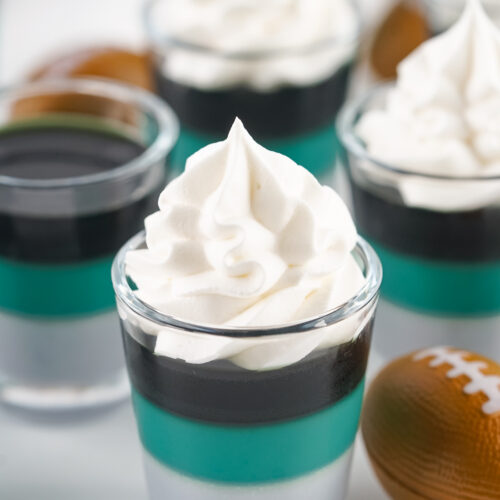 Eagles Jello Shots 24 Layered dessert shooters topped with whipped cream, featuring teal and black jelly layers in clear glasses, resemble Eagles Jello Shots, surrounded by decorative mini footballs on a white background.