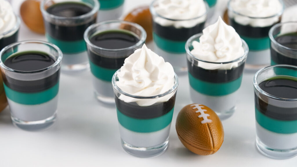 Eagles Jello Shots 2 Small layered desserts in clear glass cups with whipped cream on top are arranged alongside a miniature football, cleverly disguised as Eagles Jello Shots for an extra festive touch.
