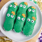 Green alligator-shaped cookies with candy eyes rest on a white plate, capturing the spirit of Mardi Gras Alligator Cookies. They are surrounded by colorful beads and bells.