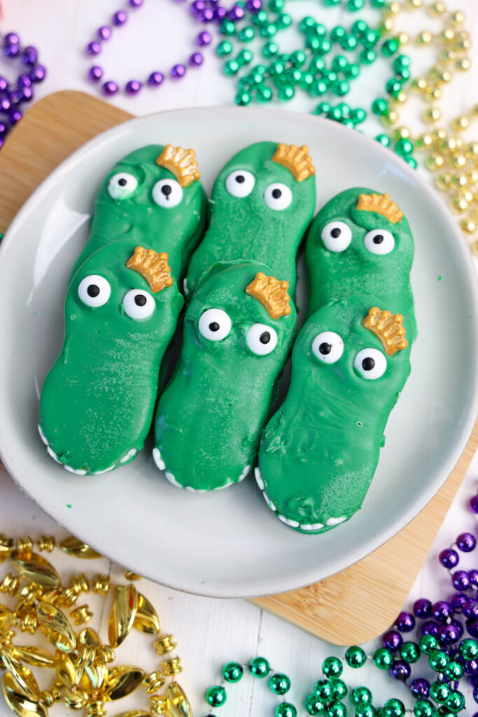 Easy Mardi Gras Alligator Cookies (No-Bake) 13 Green alligator-shaped cookies with candy eyes rest on a white plate, capturing the spirit of Mardi Gras Alligator Cookies. They are surrounded by colorful beads and bells.