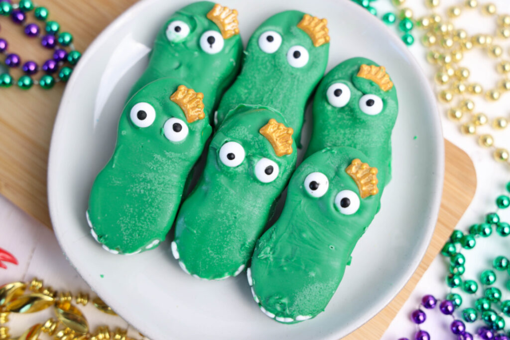Easy Mardi Gras Alligator Cookies (No-Bake) 11 Plate with six green alligator cookies featuring googly eyes and golden scales, surrounded by colorful beads on a wooden surface. These Mardi Gras Alligator Cookies bring a playful touch to any celebration.