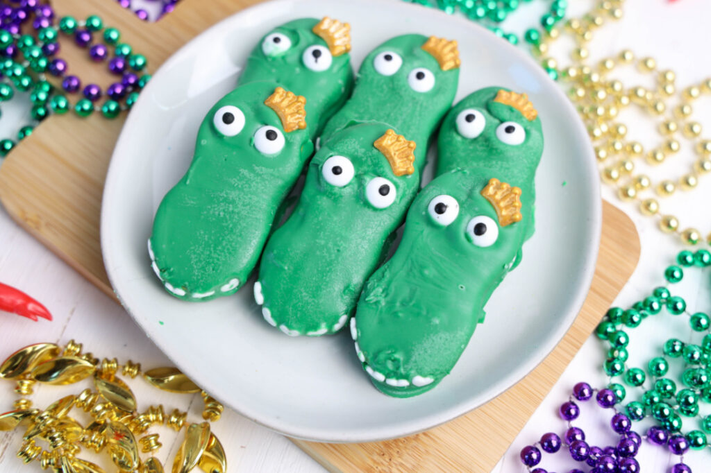 Easy Mardi Gras Alligator Cookies (No-Bake) 2 Mardi Gras Alligator Cookies in dinosaur shapes are artfully arranged on a white plate, surrounded by vibrant, colorful beads.