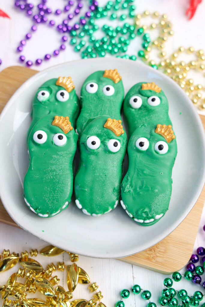 Easy Mardi Gras Alligator Cookies (No-Bake) 14 A plate of vibrant green alligator cookies, whimsically shaped like crocodiles with white eyes and shimmering gold details, surrounded by a festive array of colorful beads. Perfect for adding a playful touch to your Mardi Gras celebrations!.