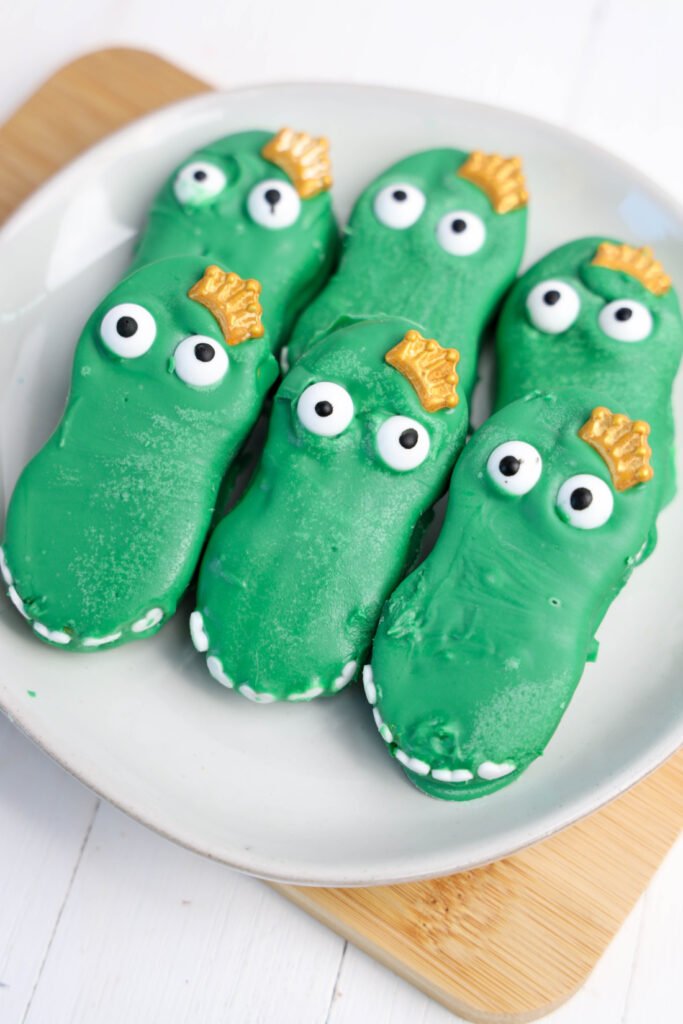 Easy Mardi Gras Alligator Cookies (No-Bake) 12 Green crocodile-shaped cookies, adorned with white and black eyes and yellow crowns, rest on a white plate. These delightful treats, resembling Mardi Gras Alligator Cookies, add a festive twist to any table setting.