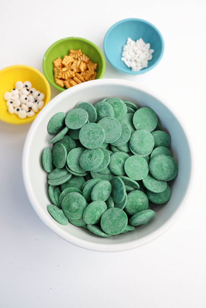 Easy Mardi Gras Alligator Cookies (No-Bake) 4 Green candy melts in a white bowl, surrounded by small bowls with white candy eyes, gold pieces, and snowflake sprinkles—perfect for crafting Mardi Gras Alligator Cookies.