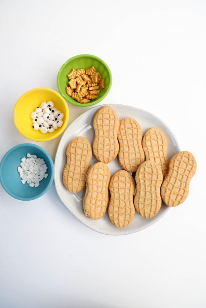 Easy Mardi Gras Alligator Cookies (No-Bake) 3 A plate of alligator-shaped cookies is surrounded by colorful bowls containing decorative candy eyes, snowflake sprinkles, and gold cereal pieces on a white background.